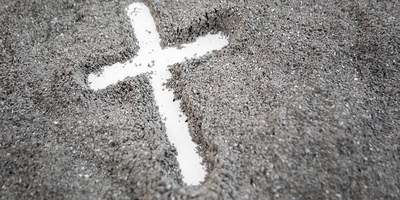 Ein mit dem Finger gezeichnetes weißes Kreuz erscheint auf grauem Asche-Hintergrund.